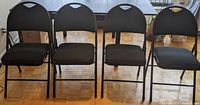 Set of four black folding chairs lined up, showing overall condition and style including seating and back padding.