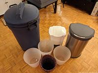 Photo showing all garbage cans and small waste bins together on a wooden floor, including Simple Human sensor can, Rubbermaid outdoor can, and 5 small bins