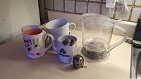 Overall view of all items on counter: 5 mugs, 1 clear David's Tea pitcher with strainer, 2 metal tea strainers