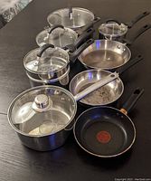 Collection of pots and pans on a dark surface including several with glass lids and black handles.