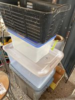 Stack of three plastic storage totes with lids: large gray tote at bottom, medium clear tote with white lid in middle, and smaller translucent white tote with blue lid on top. On top edge rests the black plastic basket