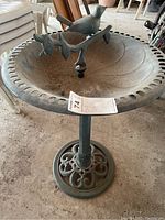 Full view of metal bird bath showing circular scalloped edge basin with bird figure and vine branch in center, mounted on single column and ornate base.