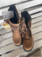 Top-down photo showing pair of brown STC boots with laces tied and paper tag attached.