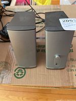 Pair of Bose Companion 2 Series II speakers shown from front and side angle on cardboard surface with cables visible in the background. One speaker with volume knob on front panel.