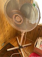 Full view of the white oscillating pedestal fan showing the fan head, protective mesh grille, and adjustable stand with cross base.