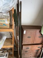 Five weathered metal fence posts leaning against a wall next to wooden furniture and shelving. Posts show rust and wear with multiple holes for fencing attachments.
