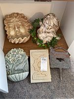 Overview of lot showing lion face plaque, cherub statue surrounded by leaves, relief wall pocket, etched concrete plaque, and metal garden sign.