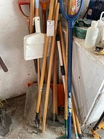 Photo showing a cluster of wooden-handled garden tools including shovels, two brooms with metal dustpans, and dustpans