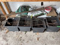 Four large black plastic planters partially filled with soil, each labeled 15x15x12 inches with green tape. Metal tomato cages and several long metal rods laid across planters using them as support. Bags of potting soil and folded paper yard bags placed behind the planters.