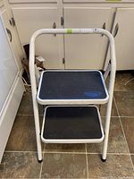 Front view of the two step stepladder showing white metal frame and black steps.