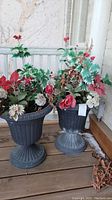 Two dark gray plastic urns with faux floral arrangements on a wooden deck against a textured wall.