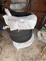 Four black radial winter tires stacked, wrapped in white plastic bags printed 'Motomaster', stored on concrete floor