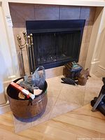 Full view of the fireplace area showing brass-handled fireplace tools on stand, large round metal container with firewood and bags, metal fish and turtle décor on floor, metal stand box and brass container near fireplace.