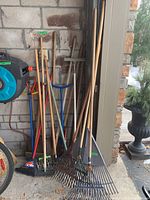 Multiple wood handle gardening tools including shovels, leaf rakes, garden rakes, broom, and hand tiller leaned against a wall