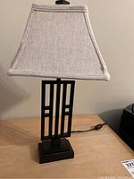 Front view of a single lamp showing black metal geometric base and beige fabric lampshade.