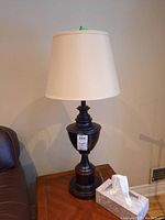 Full view of one table lamp showing the dark wood turned base and white fabric shade, placed on wooden table near leather chair.