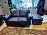 Front angled view of Natuzzi dark brown leather loveseat with matching ottoman on right side, placed in a room with hardwood floor and large window in background.