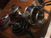 Overview of multiple pots and pans including stacked non-stick frying pans, stainless steel saucepans, and lids with black handles on wooden table.