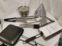 Various cooking and kitchen items arranged on white cloth including stainless steel pot, BBQ tools, wire basket with cloth liner, berry strainer, grill brush, and perforated grill basket