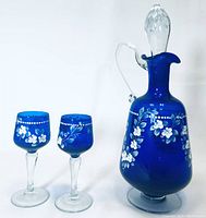 Blue glass decanter with clear stopper and handle, next to two blue glasses with hand-painted white floral designs.