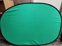 Foldable pop out green screen in oval shape with green fabric surface and black trim.