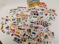 Photo showing a tin container filled with a large assortment of postage stamps spread out around it on a white surface.