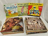 Photo showing the vintage vinyl records stacked in two piles. Top visible records include Disney Winnie the Pooh, Disney Johnny Appleseed, The Smurfs All Star Show, The Monkees, and Joan Baez albums.