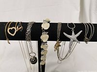 Six necklaces hanging on a black stand, showing a variety of styles and pendants: gold-tone heart, silver shell, white stone cluster, gold-tone turtle, silver starfish, and gold-tone leaf with dark stone pendants.