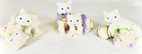 Front view of three white cat figurines, showing blue eyes and decorative bows or floral elements.