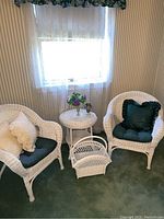 Photo showing two white wicker chairs with green seat cushions and cream decorative pillows, round white wicker table, white wicker magazine holder, silver vase with faux flowers, and plug-in lamp/nightlight on green carpet in room with striped wallpaper and lace curtains.