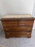 Front view of 3-drawer wooden dresser with brass knobs and pulls, crocheted doily on top.