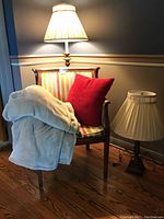 Wood and fabric armchair with striped upholstery, red pillow, and cream throw blanket placed with floor and table lamps, all items shown in a corner against a blue wall with chair rail, hardwood floor.