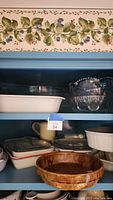 Photo showing a mix of white and brown ceramic baking dishes and glass serving bowls with decorative floral pattern on the top shelf of a blue cabinet.
