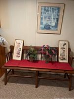 Full view of wooden bench with red cushion, two small framed pieces on bench, and decorative plants, with large framed pretzel cart artwork hanging on wall above.
