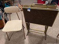 Beige folding chair next to stacked folding TV trays with brown patterned tops and gold frames