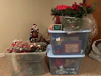 Three plastic bins with assorted Christmas decorations including gift bags, cards, greenery, wreath on top, Santa and snowman figurines partially visible
