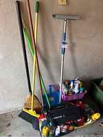 Full view of cleaning tools including brooms, mop, squeegee, blue dustpan, plastic basket with various cleaning liquids, and Oreck XL vacuum at base.
