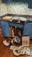 Wide view of kitchen counter and floor showing mix of kitchen items: cutting boards, mixing bowl, cooking pans, and serving trays.