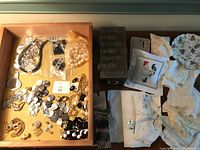 First photo: Drawer with assorted costume jewelry, embroidered handkerchiefs folded and stacked, heavy engraved metal box to right.