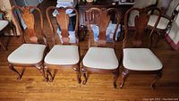 Four wooden dining chairs with carved backs and cream upholstered seats arranged in a row on hardwood floor.
