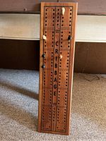Wooden cribbage board with pegs inserted for game play, labeled 'CRIBBAGE' and divided into columns with drilled holes.