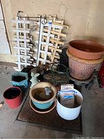 Photo showing several white wooden garden fencing pieces, assorted flower pots in different materials and colors, a metal plant hanger, and garden statues including angel bust and bird statue.