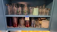 Glass cabinet shelf with clear glasses, plastic cups in purple and frosted white, and stacked ceramic coasters.