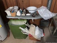 Various household and laundry items on an ironing board including small iron, white enamel basin, measuring tape, and green woven hamper next to board