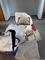 Photo showing assorted bedding including pillows, blankets, purple floral coverlet, and laundry basket with folded bedding items.