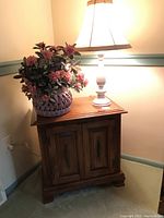 Photo showing one wooden nightstand with two cabinet doors, white lamp on top with fabric shade, and faux pink flowering plant in basket.