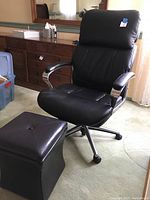 Main image showing the black adjustable office chair with padded armrests and the black square storage ottoman beside it on a carpeted floor.