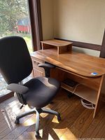 Office desk with attached small elevated wood shelf and black adjustable chair in front of window.