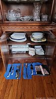 Photo showing china serving platters and bowls on middle shelf with silver utensils and serving tools on blue cloths on floor in front of cabinet