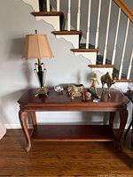 Wooden sofa table with multiple collectibles including lamp, statues, and figurines on top. Table is positioned in front of staircase with hardwood flooring beneath.